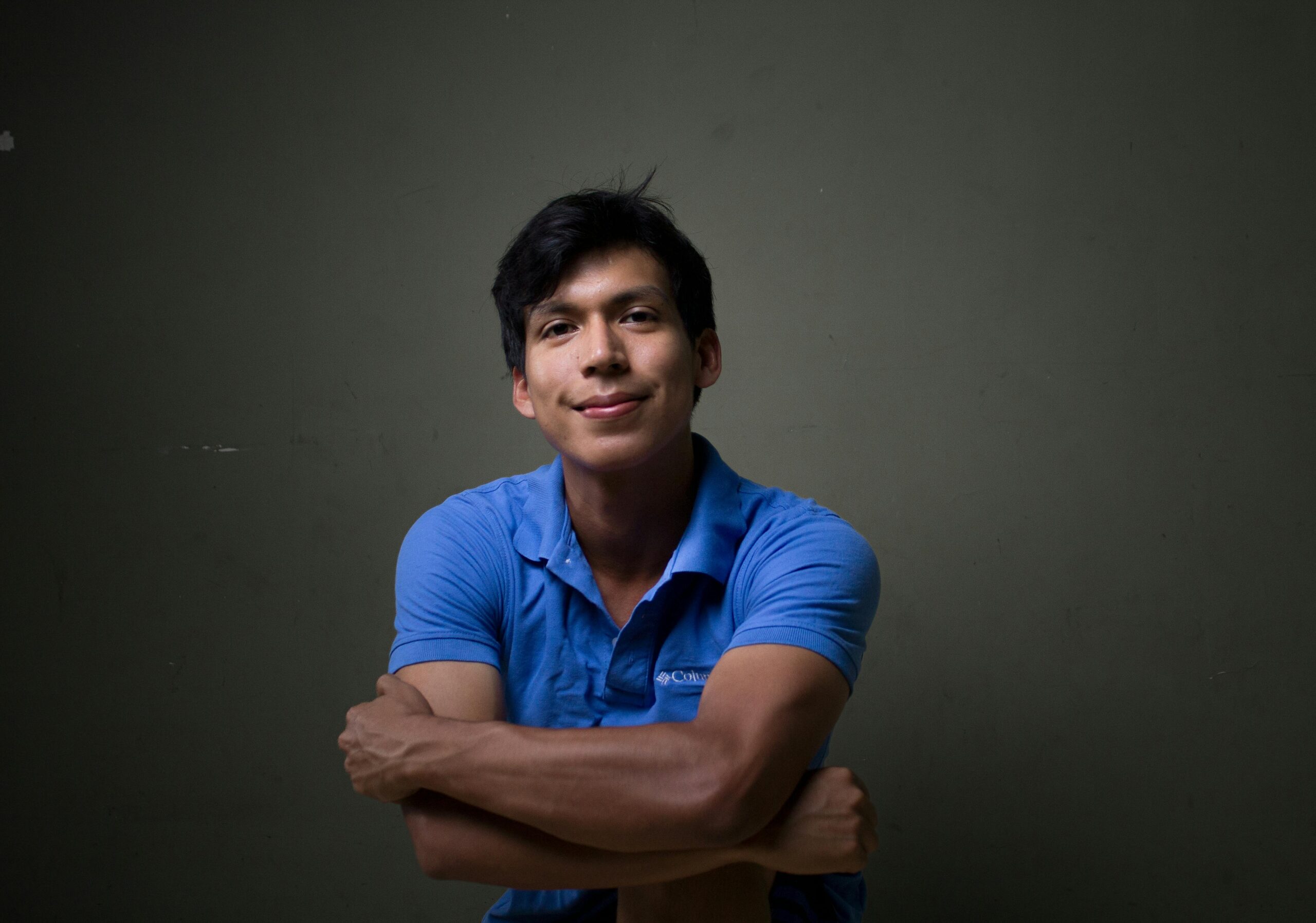 Young Hispanic man smiling confidently in a blue shirt against a dark background.